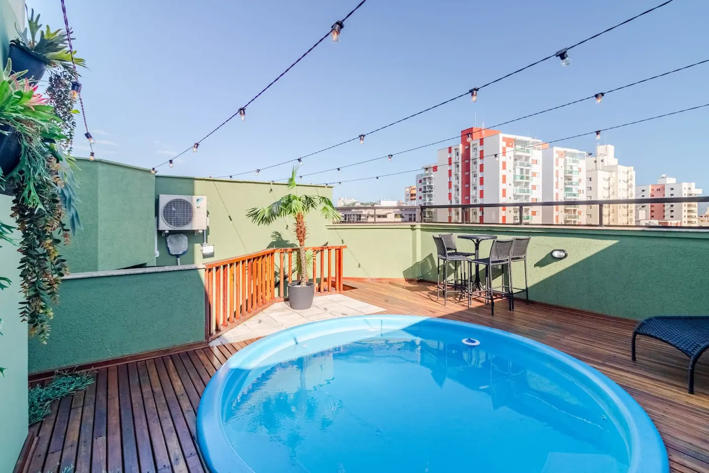 Terraço do duplex no Guarujá com piscina privativa, deck de madeira e varal de luzes, num dia de céu azul